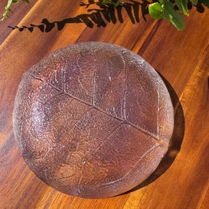 May include: A clear, round glass dish with a leaf design. The dish has a textured surface and is brown in color. The dish is sitting on a wooden surface.
