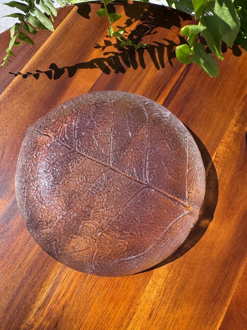 May include: A clear, round glass dish with a leaf design. The dish has a textured surface and is brown in color. The dish is sitting on a wooden surface.