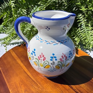 May include: White ceramic jug with a blue handle and rim, decorated with colourful floral patterns in red, blue, and yellow. The jug is placed on a wooden surface, displaying a classic design for serving drinks.