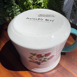 May include: White ceramic mug with a teal handle and a strawberry design. The mug has a ribbed texture and the text "Nathalie Lete for Anthropologie" is printed on the bottom. Dishwasher and microwave safe.