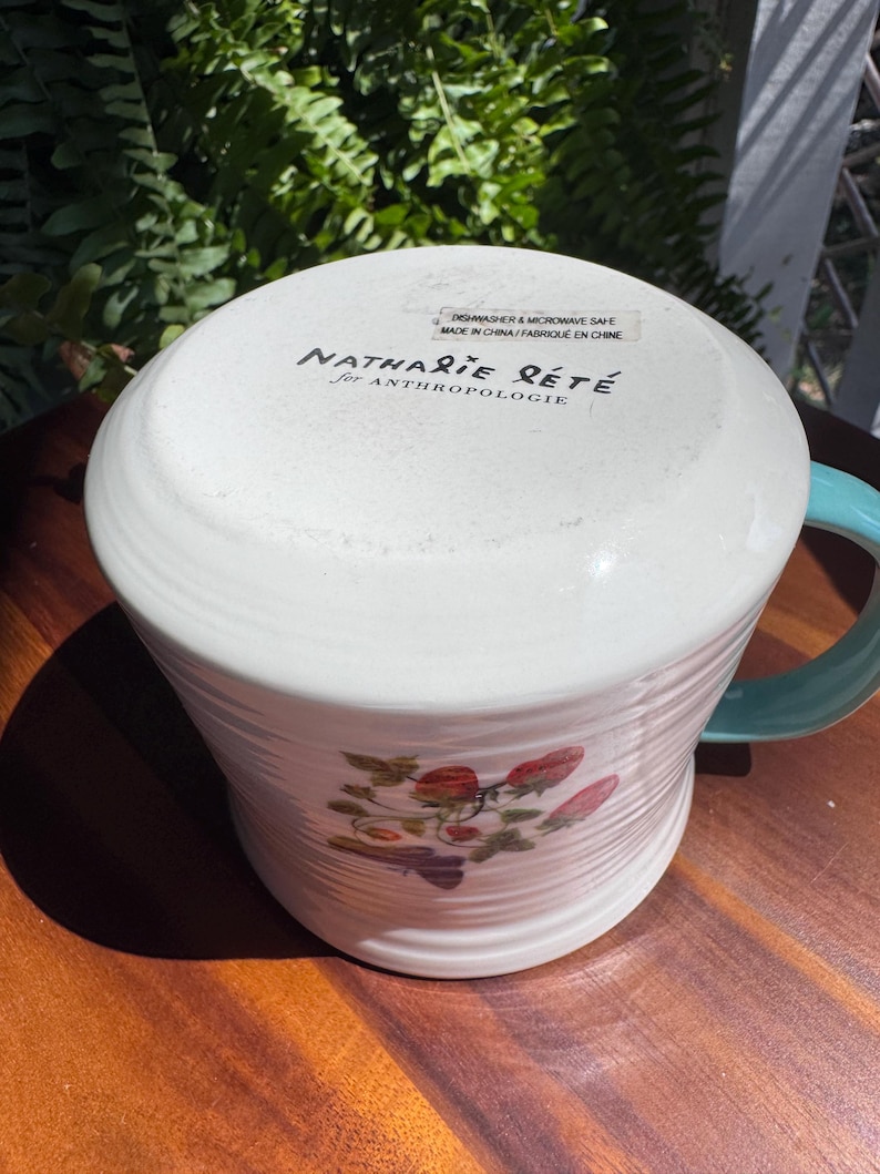 May include: White ceramic mug with a teal handle and a strawberry design. The mug has a ribbed texture and the text "Nathalie Lete for Anthropologie" is printed on the bottom. Dishwasher and microwave safe.