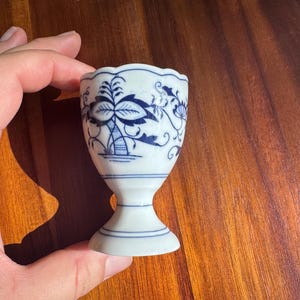 May include: A white porcelain egg cup featuring a scalloped rim and a blue floral pattern. The base is pedestal-style with thin blue horizontal lines encircling the stem and the foot of the cup, providing a stable foundation.