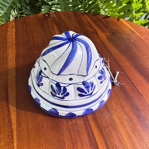 Könnte beinhalten: Eine Keramikform mit weißer Basis und handgemalten blauen Blumen- und Wirbelmustern. Das kuppelförmige Objekt zeigt ein spiralförmiges Design an der Oberseite, eine Reihe blauer Punkte und ein wiederkehrendes Blütenblattmotiv am unteren Rand.