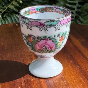May include: A white porcelain egg cup featuring a hand-painted floral design with prominent pink, green, and orange accents. The cup has a flared rim, a rounded bowl, and a sturdy pedestal base, decorated with intricate patterns around the top.