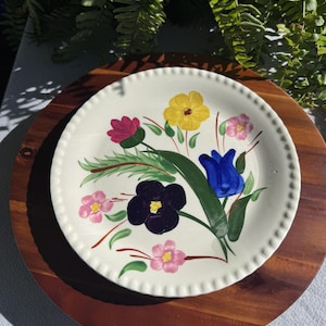 May include: A round, off-white ceramic plate featuring a scalloped edge and a hand-painted floral design. The center displays a bouquet of colorful flowers including a large dark purple bloom, a bright yellow flower, a blue tulip, and pink blossoms.