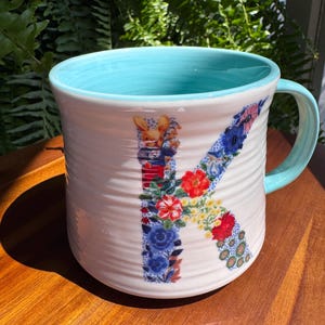 May include: White ceramic mug with a light blue interior and handle. The letter "K" is decorated with a floral pattern in red, blue, and yellow. The mug has a textured, ridged exterior and sits on a wooden surface.