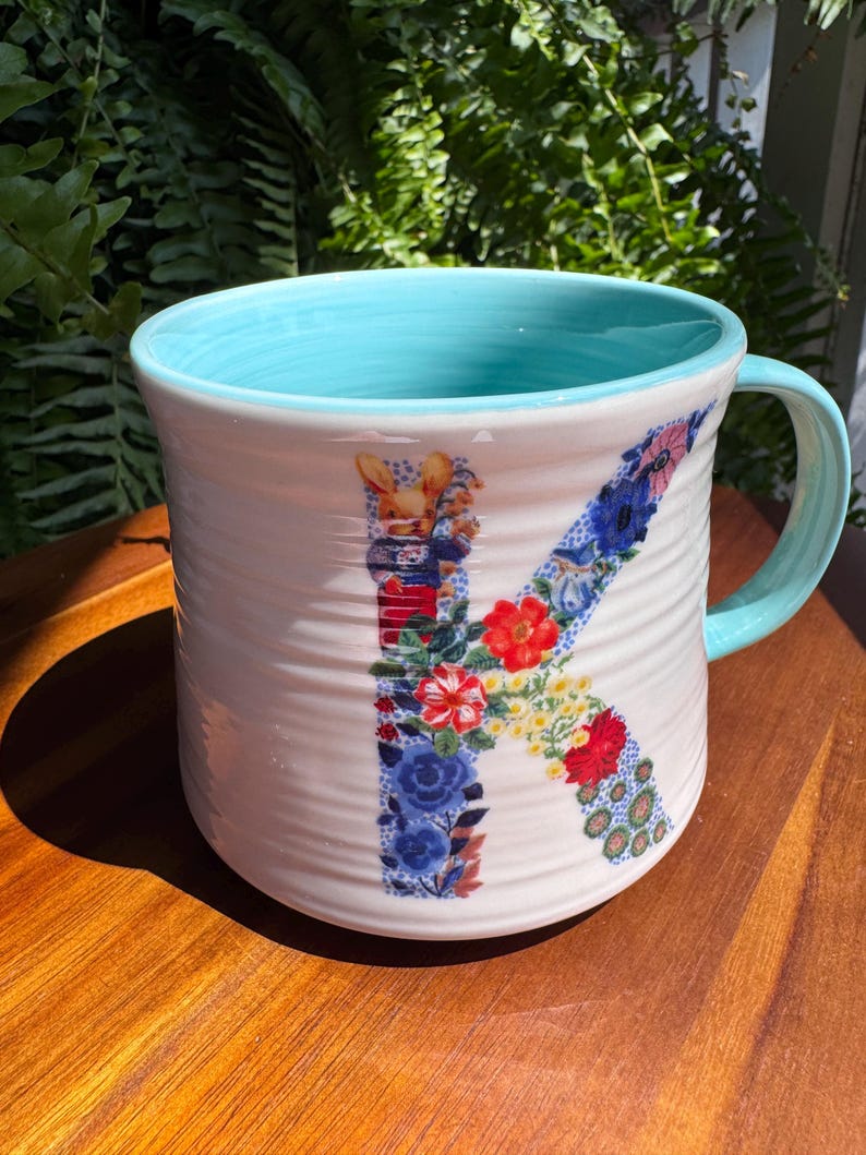 May include: White ceramic mug with a light blue interior and handle. The letter "K" is decorated with a floral pattern in red, blue, and yellow. The mug has a textured, ridged exterior and sits on a wooden surface.