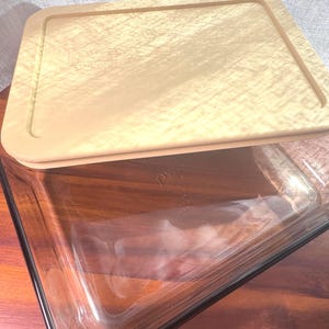 May include: A clear Pyrex rectangular baking dish with a tan lid. The lid has the word "Pyrex" printed on it. The dish is sitting on a wooden surface. The lid is slightly ajar, revealing the interior of the dish.