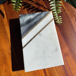 May include: A rectangular marble board featuring a split design with a dark grey, veined section and a larger white section. Two thin, parallel gold-toned metal strips run diagonally across the surface, separating the two stone colors.