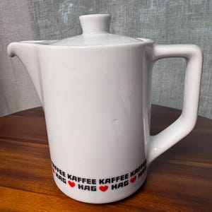 May include: White ceramic coffee pot with a lid and handle. The pot has a spout for pouring and a black and red design that reads "KAFFEE HAG" with red hearts. The pot is on a wooden surface.