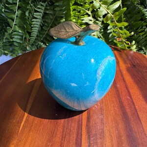 May include: A decorative blue apple sculpture with a brass leaf and stem. The apple has a glossy finish and sits on a wooden surface. The sculpture is a unique home decor item.
