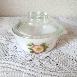 May include: A white casserole dish with a clear glass lid. The dish is decorated with a floral pattern featuring a pink and yellow flower and green leaves. It has two small handles and is likely made of ceramic or glass.