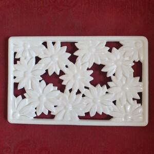 May include: A white rectangular decorative piece with a floral design. The piece features a series of daisy-like flowers arranged within a rectangular frame. The flowers are raised, creating a textured surface.