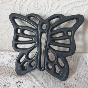May include: A dark grey butterfly-shaped trivet. The metal trivet features an openwork design, with the butterfly's wings and body clearly defined. The cut-out patterns create a detailed appearance. It is designed to protect surfaces from hot items.