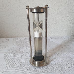 May include: A silver-toned metal hourglass featuring a clear glass cylinder containing white sand. The frame consists of a circular top and base connected by three vertical pillars, with a reflective metallic finish throughout the structure.