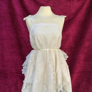 May include: Ivory-colored lace dress with a square neckline and spaghetti straps. The dress features a floral lace overlay and a layered skirt with a handkerchief hemline. A matching ribbon belt cinches the waist.