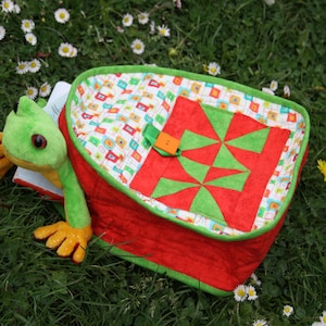Może przedstawiać: Czerwono-zielony pikowany plecak z pluszową zabawką w kształcie żaby. Plecak ma małą kieszeń z przodu z zielono-czerwonym wzorem patchworkowym.