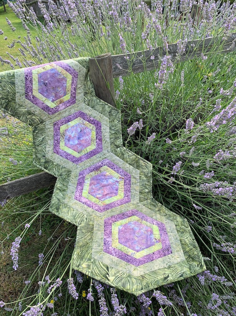 May include: A green and purple quilted table runner with a hexagonal pattern. The runner is made of fabric with a floral print and is lying on a bed of lavender.