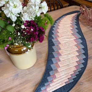 May include: A blue and pink quilted table runner with a curved shape. The runner is made of fabric squares sewn together in a pattern. The runner is on a wooden table.
