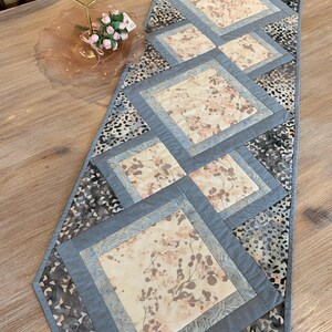 Op de afbeelding: Een grijze en beige tafelrunner met een geometrisch patroon van vierkanten. De vierkanten zijn gemaakt van verschillende stoffen met bloemenpatronen. De runner is ongeveer 40 cm breed en 122 cm lang.