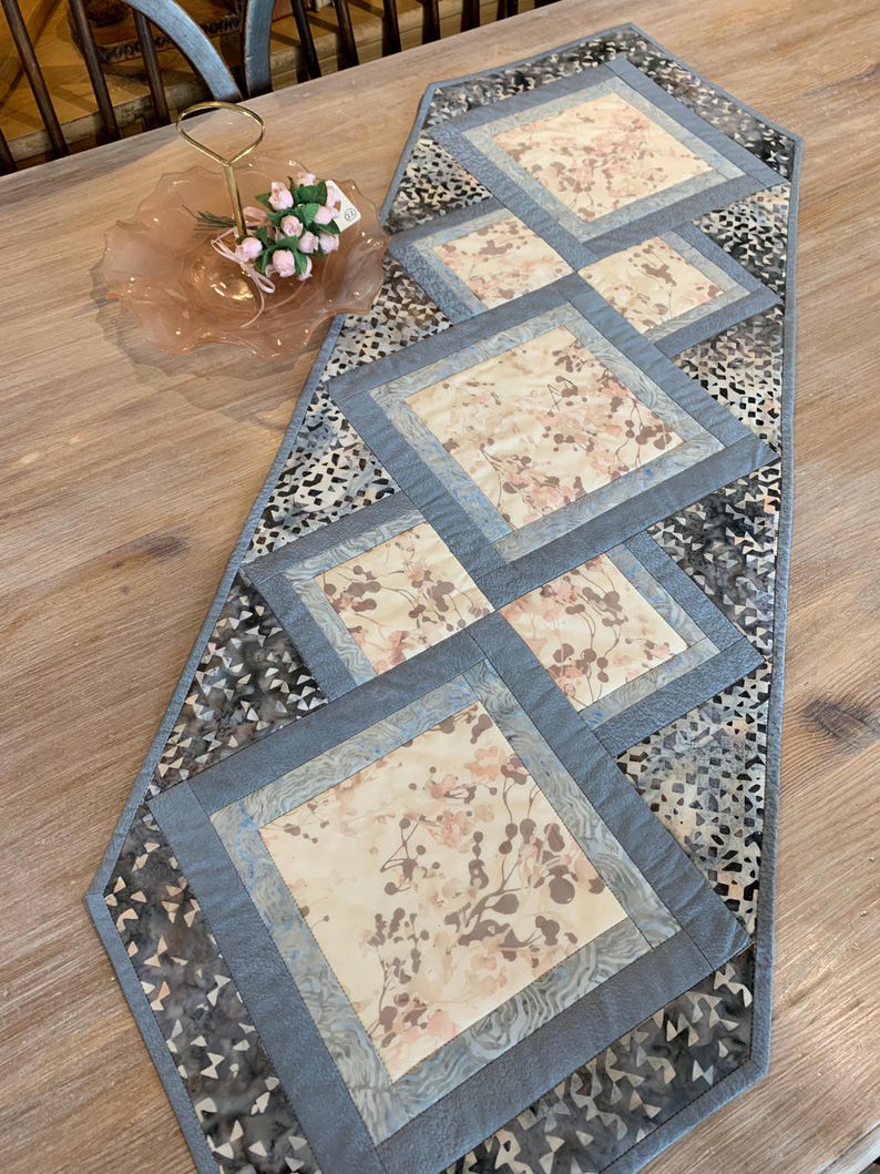 Op de afbeelding: Een grijze en beige tafelrunner met een geometrisch patroon van vierkanten. De vierkanten zijn gemaakt van verschillende stoffen met bloemenpatronen. De runner is ongeveer 40 cm breed en 122 cm lang.