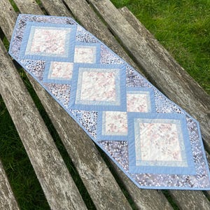 Op de afbeelding: Een blauwe en witte gestikte tafelrunner met een geometrisch patroon. De runner is gemaakt van stof met een bloemenprint en ligt op een houten oppervlak.