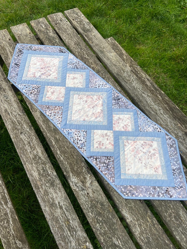 Op de afbeelding: Een blauwe en witte gestikte tafelrunner met een geometrisch patroon. De runner is gemaakt van stof met een bloemenprint en ligt op een houten oppervlak.