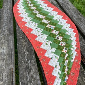 May include: A red, green, and white quilted table runner with a Christmas theme. The runner is made of patchwork squares with various Christmas-themed prints. The runner is curved and has a scalloped edge.