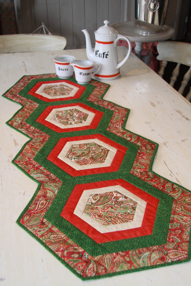 May include: A green, red and cream quilted table runner with a paisley pattern. The runner is made up of six hexagonal shapes.