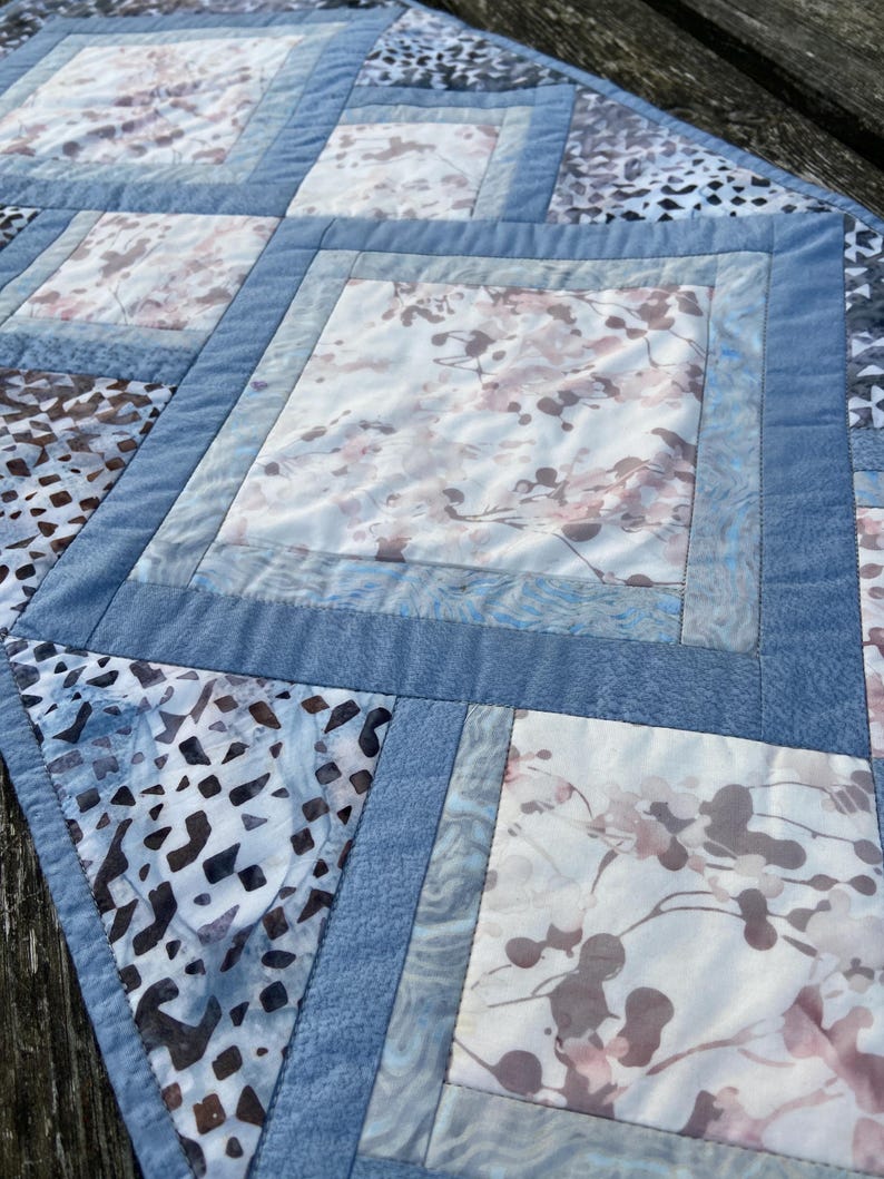 Op de afbeelding: Een blauwe en witte gestikte tafelrunner met een geometrisch patroon. De runner is gemaakt van verschillende stoffen met een verscheidenheid aan patronen, waaronder bloemen- en geometrische ontwerpen.