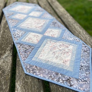 May include: A blue and white quilted table runner with a diamond pattern. The runner features a floral print and a light blue border.