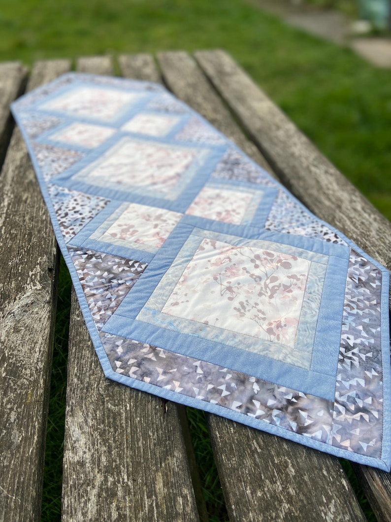 Op de afbeelding: Een blauwe en witte gestikte tafelrunner met een ruitpatroon. De runner heeft een bloemenprint en een lichtblauwe rand.