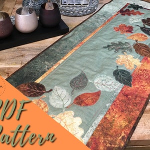 May include: A PDF pattern for a table runner featuring a design of autumn leaves in shades of brown, orange, and green. The runner is on a wooden table with candlesticks and candles.