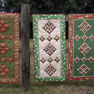 May include: Three quilted table runners with a diamond pattern in red, green and white. The runners are hanging on a wooden fence.