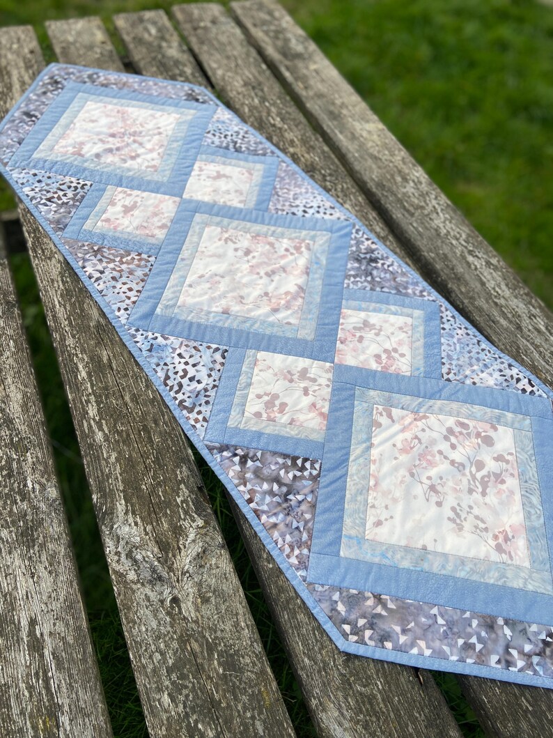 Op de afbeelding: Een blauwe en witte tafelrunner met een ruitpatroon. De runner is gemaakt van stof met een bloemenprint en is gestikt.