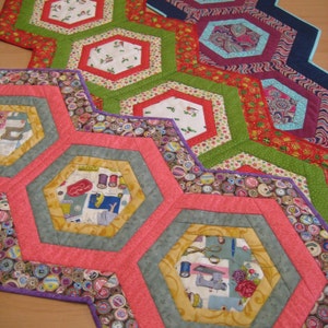 May include: A set of five hexagonal table runners featuring a patchwork design. The runners are crafted from a variety of fabrics with diverse patterns and colours, including red, green, blue, and purple. The runners are layered on top of each other, creating a visually appealing and intricate design.