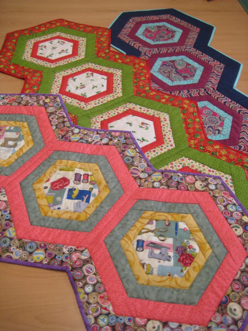 May include: A set of five hexagonal table runners featuring a patchwork design. The runners are crafted from a variety of fabrics with diverse patterns and colours, including red, green, blue, and purple. The runners are layered on top of each other, creating a visually appealing and intricate design.