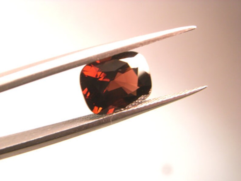 May include: A dark red gemstone with facets, held by tweezers against a white background.