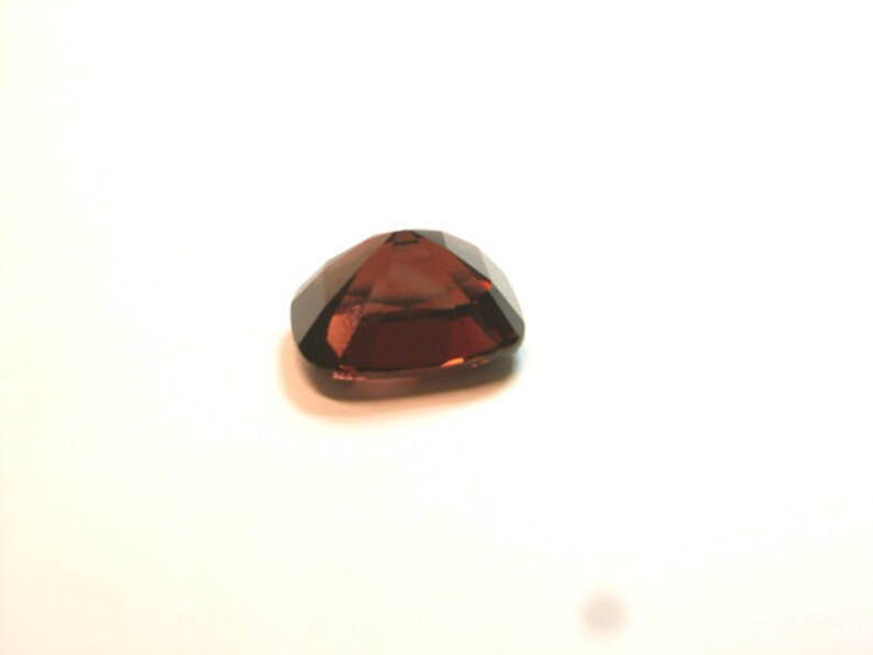 May include: A dark red gemstone with a faceted cut. The gemstone is set in a square shape.