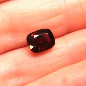 May include: A deep red gemstone with a cushion cut. The gemstone is set in a ring.