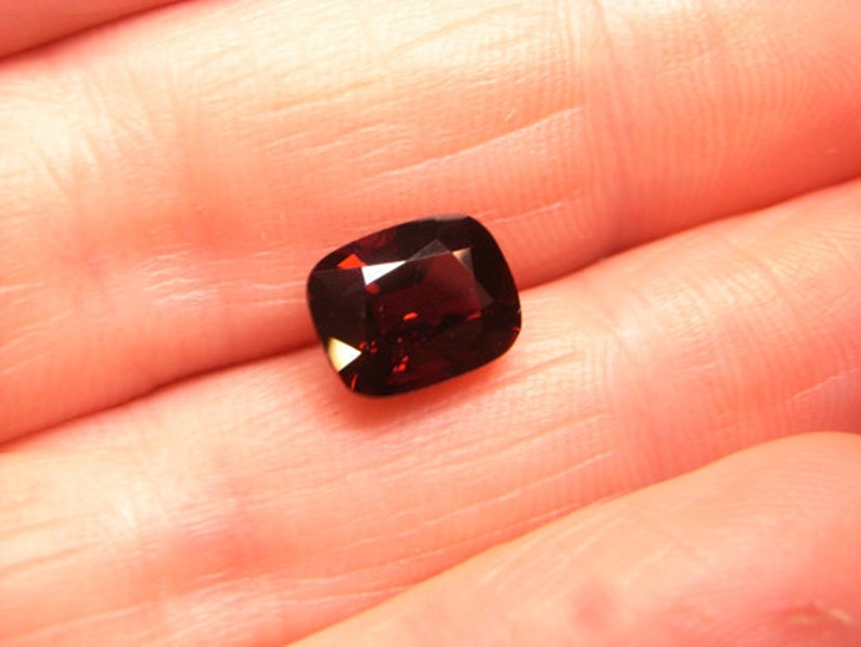 May include: A deep red gemstone with a cushion cut. The gemstone is set in a ring.