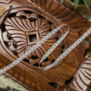 May include: A white crochet ribbon with a delicate pattern is displayed on a wooden tray with intricate carvings. A crochet hook is also visible in the image.