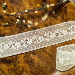 May include: White lace trim with a floral pattern and small dots. The trim is on a wooden surface.