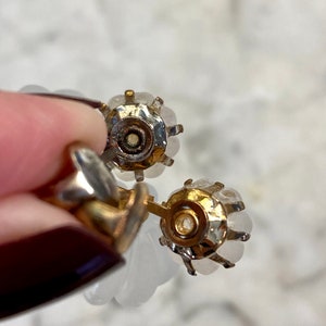Puede incluir: Un par de pendientes de tono dorado con piedras blancas nacaradas. Los pendientes tienen forma redonda y est&aacute;n engastados con varias piedras.