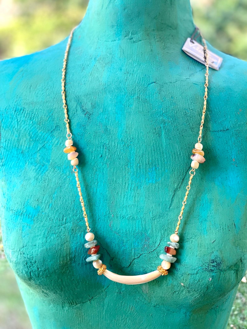 Puede incluir: Un collar de cadena dorada con un colgante curvo blanco y cuentas de piedras preciosas multicolores. Las cuentas son en tonos de marr&oacute;n, verde, rojo y blanco.