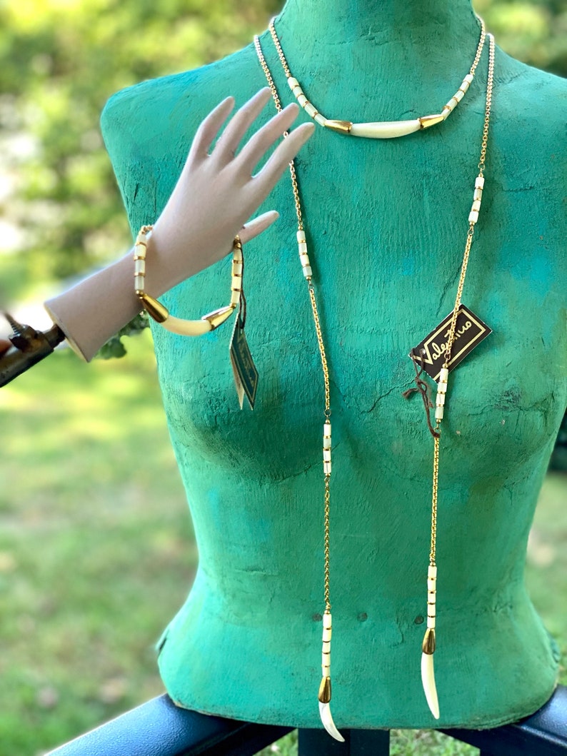 Puede incluir: Un collar y un brazalete de cadena dorada con cuentas blancas de hueso y detalles dorados. El collar tiene dos cadenas largas con colgantes de hueso en los extremos. El brazalete es una sola cadena con un colgante de hueso. El conjunto se muestra en un maniqu&iacute; verde.