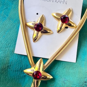 May include: A gold chain necklace with a red jeweled star pendant and matching earrings. The jewelry is on a white card with the text "ROBIN KAHN".
