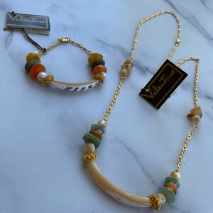 Puede incluir: Un conjunto de collar y pulsera de oro con un colgante de concha y cuentas verdes, naranjas y blancas. El conjunto viene con dos etiquetas que dicen "Valentino".