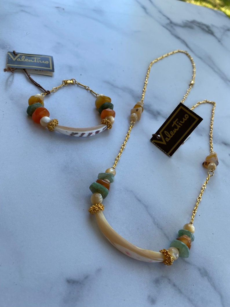 Puede incluir: Un conjunto de collar y pulsera de oro con un colgante de concha y cuentas verdes, naranjas y blancas. El conjunto viene con dos etiquetas que dicen "Valentino".