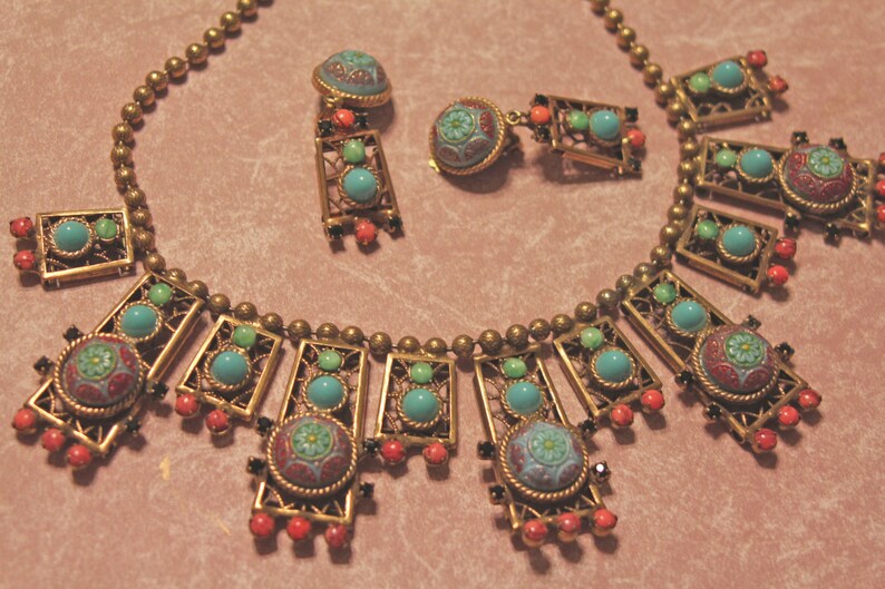 Puede incluir: Un collar de oro con detalles en turquesa y esmalte rojo. El collar presenta un dise&ntilde;o geom&eacute;trico con m&uacute;ltiples marcos rectangulares. El collar se combina con pendientes a juego.